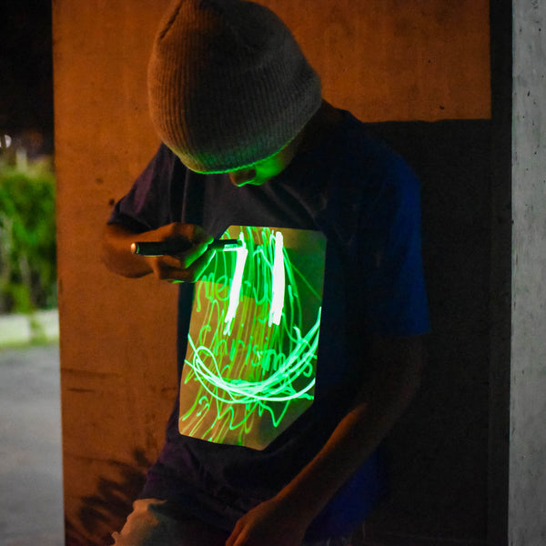 niño con camiseta azul dibujando con linterna UV sobre panel fosforescente verde
