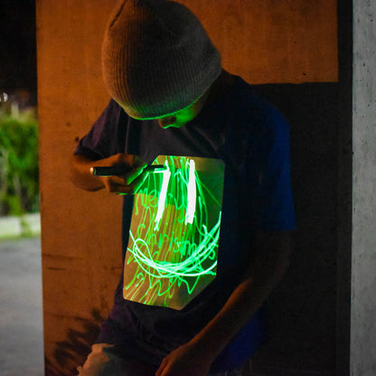niño con camiseta azul dibujando con linterna UV sobre panel fosforescente verde
