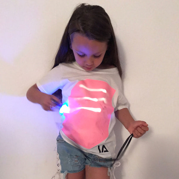 niña con camiseta blanca dibujando con linterna UV sobre panel fosforescente rosa
