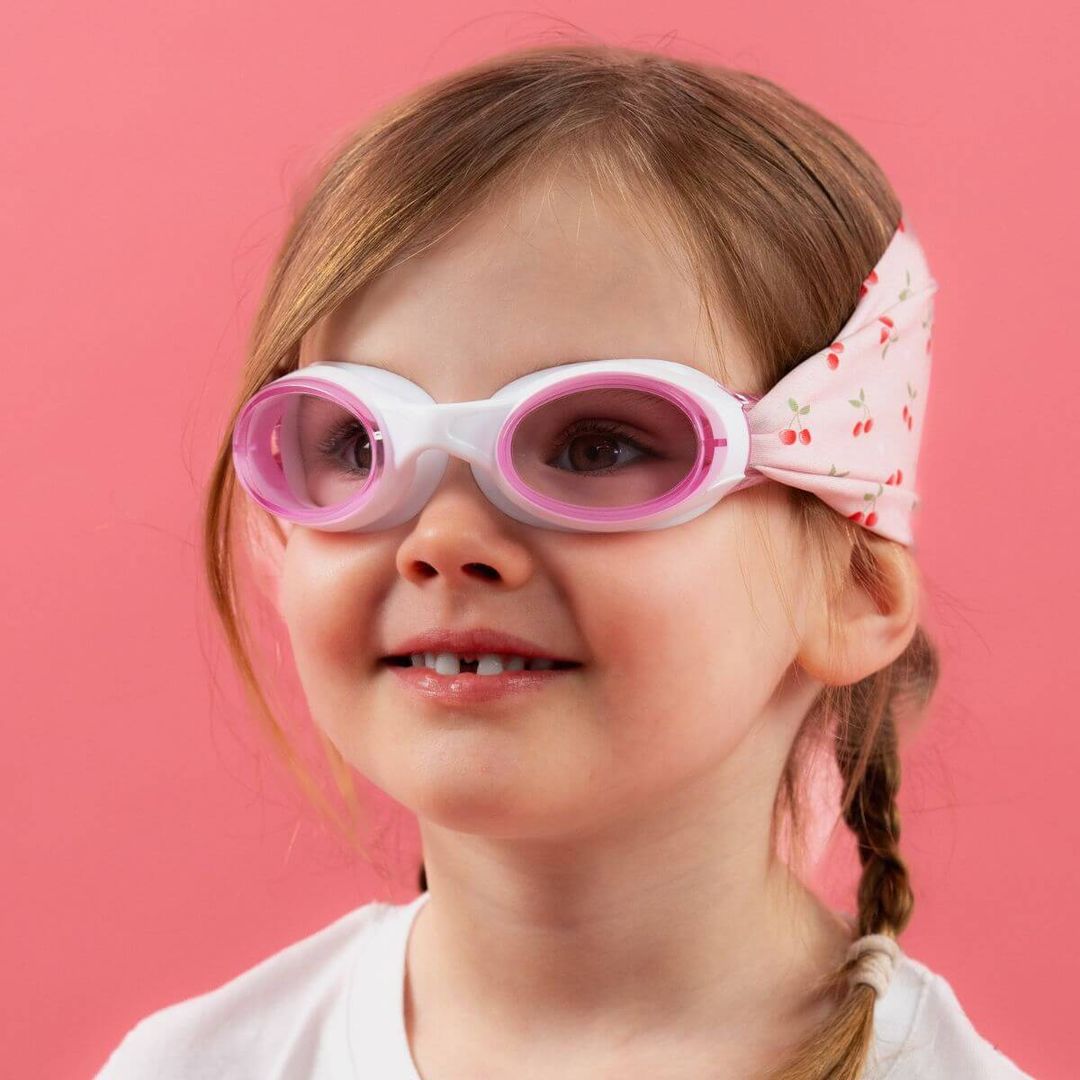 Smiling girl wears white and pink kids swim goggles with a cherry print headband.