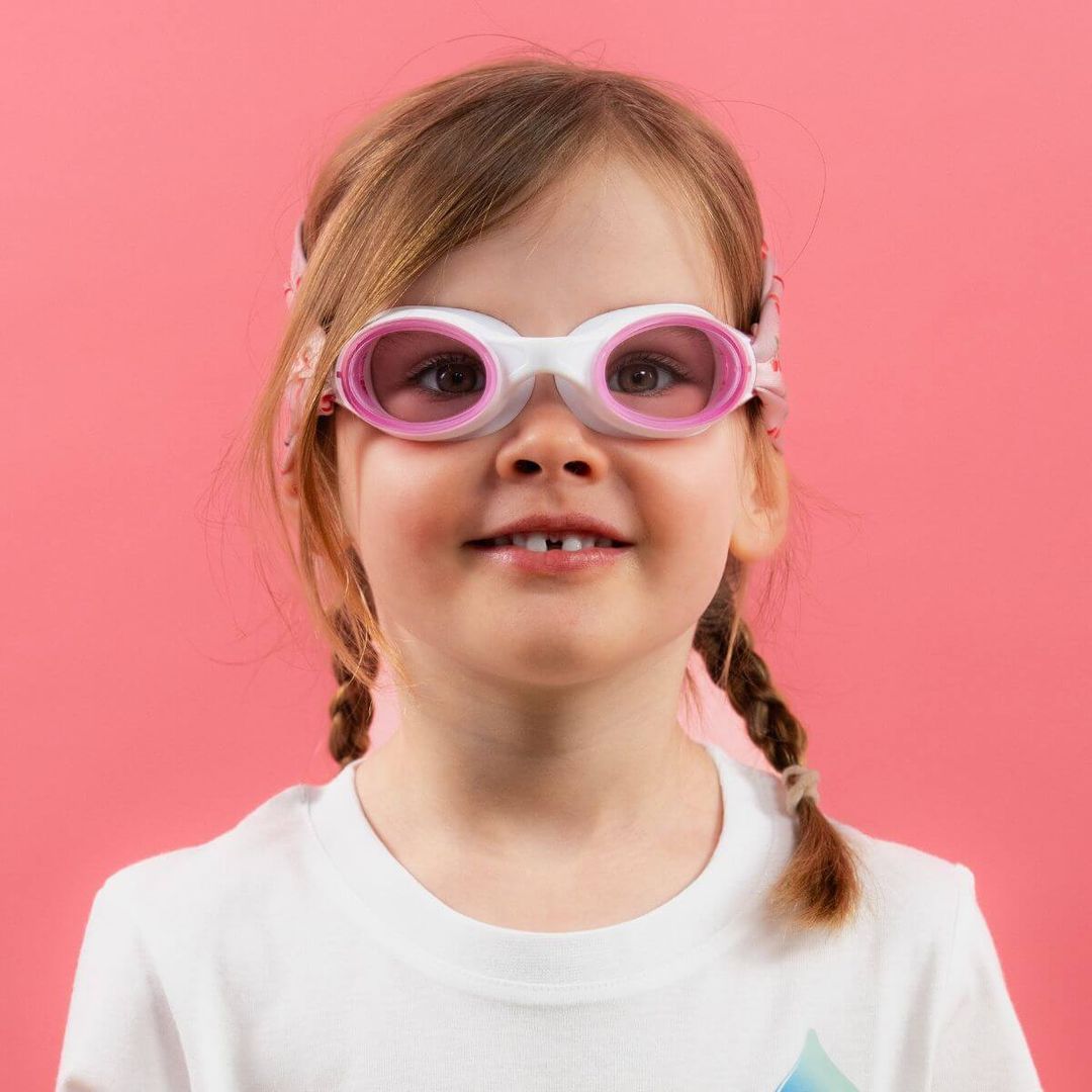 Smiling young girl wearing white and pink swimming goggles against a pink background.