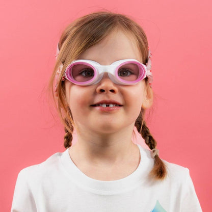 Smiling young girl wearing white and pink swimming goggles against a pink background.